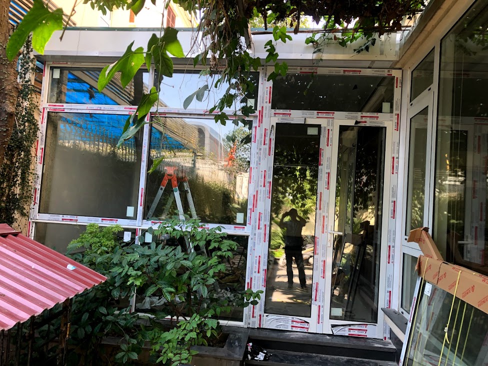 Cửa nhôm kính đang thi công tại nhà vườn, khung trắng dán tem bảo vệ, xung quanh nhiều cây xanh